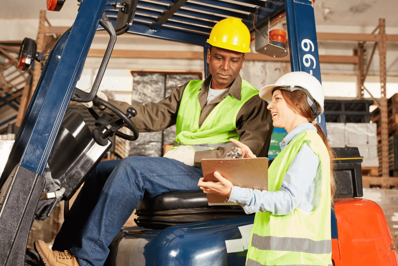 Two construction workers discussing in a forklift, both wearing helmets and safety vests.